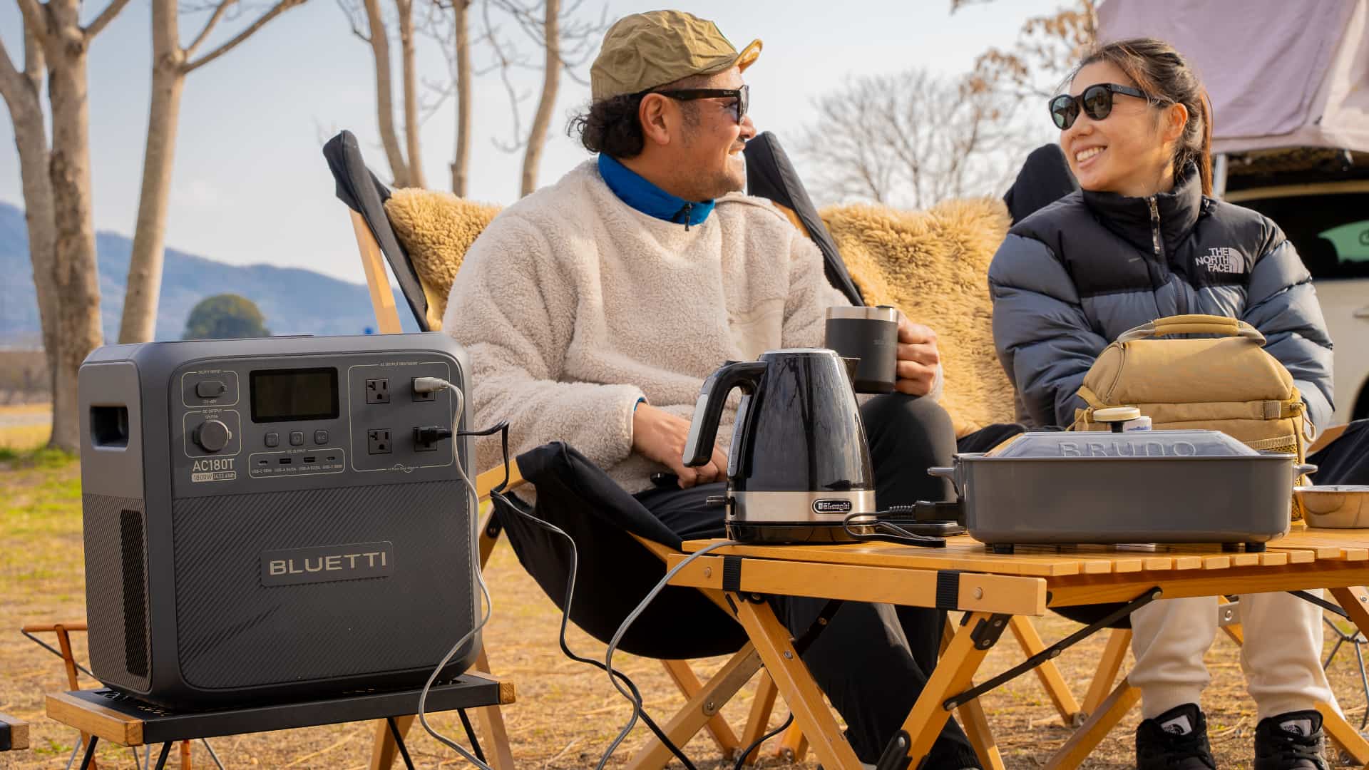 A couple sitting on a chair in the wilderness and drinking coffee, with a coffee maker plugged into Bluetti's SwapSolar AC180T power station 